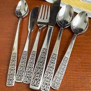 May include: A set of six silver-colored flatware pieces, including spoons, a butter knife, a fork, and a dinner knife. The handles feature a repeating decorative pattern. The silverware is arranged on a wooden surface.