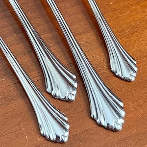 May include: Close-up of four silver-colored spoons. The spoons have a decorative, fan-like design at the base of the handle. The spoons are arranged diagonally on a wooden surface.