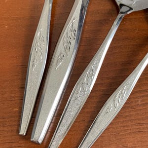 May include: Close-up of several pieces of silverware with a leaf design. The silverware is made of shiny, polished metal and is arranged on a wooden surface. The spoons and forks have a textured handle with a detailed leaf pattern.