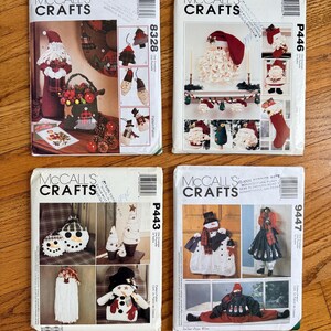May include: Four vintage McCall's Crafts pattern booklets featuring Christmas-themed crochet projects. The covers display images of Santa Claus, snowmen, Christmas stockings, and other holiday decorations. The booklets are on a wooden surface.