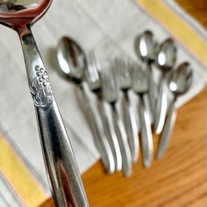 May include: Close-up of a stainless steel spoon with a decorative floral design on the handle. Several forks and spoons are in the background, arranged on a striped white and yellow cloth. The silverware is on a wooden surface.