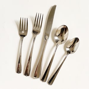May include: A set of stainless steel silverware including two forks, a knife, and two spoons. The silverware has a simple, elegant design with a polished finish, reflecting light. The forks have three and four tines, and the spoons are oval-shaped.