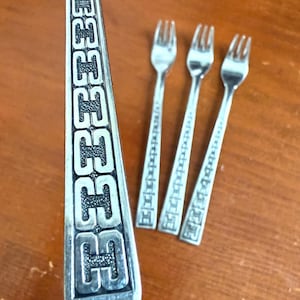 May include: Silverware set featuring a serving spoon and three small forks. The serving spoon has a decorative handle with a repeating pattern. The forks have a similar design on their handles. The silverware is displayed on a wooden surface.