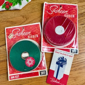 May include: Two packages of Sasheen brand ribbon, one green and one red, each labeled 300 feet, alongside a small instructional card titled How to make your Christmas gifts almost too pretty to open, featuring a graphic of a wrapped gift box.