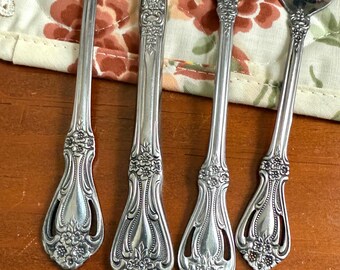 May include: Close-up of antique silverware set, including a fork, knife, and two spoons. The silver utensils have ornate floral detailing on the handles. The silverware is arranged on a floral patterned placemat and a wooden surface.