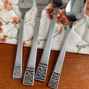 May include: Close-up of silverware set including forks and spoons. The silverware has a silver finish with a decorative design at the base of the handles. The silverware is set on a floral patterned cloth.