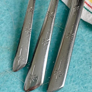 May include: Close-up of three vintage silver-colored spoons. Each spoon features a starburst design on the handle. The spoons are arranged on a light blue surface, with a yellow and white striped cloth in the background.