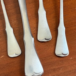 May include: Close-up of several pieces of silverware with a shiny, silver finish. The utensils include a knife and several butter spreaders, all with a decorative, heart-shaped design at the base of the handle. The silverware is arranged on a wooden surface.