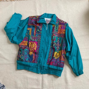 May include: A teal blue jacket with a zipper closure and a colorful patterned front. The pattern features geometric shapes and floral designs in shades of pink, yellow, purple, and green.