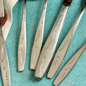 May include: Close-up of a set of silver silverware with a star and curved line design. The set includes forks and knives, arranged on a light blue surface. A patterned napkin is visible in the background.
