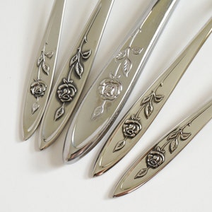 May include: Five silver forks with rose designs on the handles. The forks are arranged in a row, with the handles pointing towards the right side of the image.
