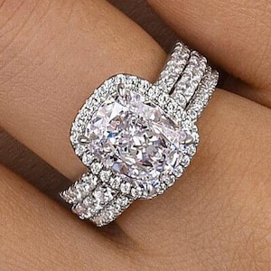 Puede incluir: Un conjunto de anillos de compromiso brillantes con una gran piedra central de diamante talla cojín rodeada por un halo de diamantes más pequeños. El anillo se combina con dos bandas de diamantes, creando un aspecto en capas. El anillo está engastado en metal blanco.