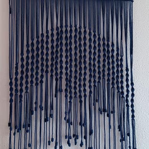 May include: A navy blue macramé wall hanging. The handcrafted piece features rows of knotted cords, creating a textured, fringed design. The wall decor is suspended from a metal rod, adding a touch of bohemian style to any space.
