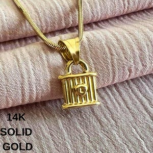 Könnte beinhalten: Halskette mit einem goldfarbenen Vorhängeschloss-Anhänger. Der Anhänger ist eine detaillierte Miniatur eines Vorhängeschlosses mit vertikalen Stäben und einer abgerundeten Oberseite. Die Kette ist dünn und goldfarben. Der Text "14K SOLID GOLD" ist sichtbar.