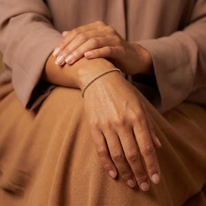 Op de afbeelding: Een delicate gouden armband bestaande uit een doorlopende streng van kleine, uniforme, gepolijste bolvormige kralen. De armband heeft een minimalistisch ontwerp met een reflecterende metalen afwerking en vormt een eenvoudige cirkel.