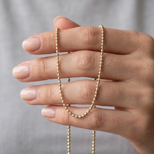 May include: A gold chain necklace featuring a series of small, round, textured beads linked together in a continuous strand. The metallic surface of the beads reflects light, creating a subtle sparkle along the length of the delicate jewelry piece.