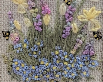 May include: A close-up of an embroidered picture of a bouquet of yellow daffodils, purple and pink flowers, and blue forget-me-nots with two bees.