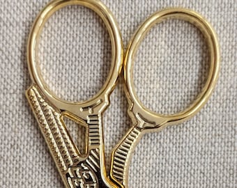 May include: Gold-toned embroidery scissors with a bird design on the handle. The scissors have a pointed tip and a small screw on the handle.