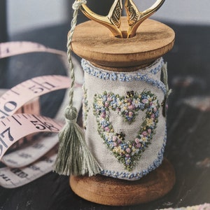 Könnte beinhalten: Eine Holzspule mit einem gestickten Herzmotiv in Blau-, Rosa- und Grüntönen. Eine goldene Schere sitzt auf der Spule. Ein grüner Quast hängt an der Spule.
