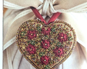 May include: A heart-shaped ornament with a gold beaded border and a red ribbon. The ornament is decorated with red fabric flowers, green accents, and gold sequins. The background features a beige bow.