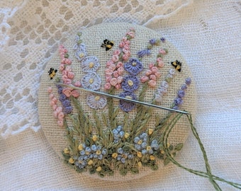 May include: A close-up of a hand-stitched embroidery hoop featuring a floral design with pink, purple, and blue flowers and green leaves. Two small yellow bees are embroidered on the hoop.