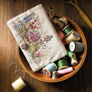 May include: A wooden bowl filled with spools of thread in various colours, including green, pink, gold, and grey. A white linen pouch with an embroidered floral design sits in the bowl. A measuring tape and a pair of scissors are also visible.