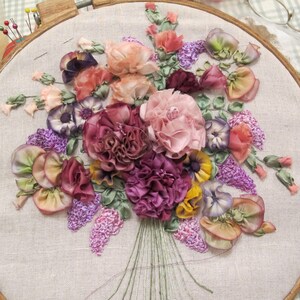 May include: A close-up of a hand-stitched embroidery hoop featuring a colorful bouquet of silk ribbon flowers. The flowers are in shades of pink, purple, yellow, and green. The embroidery is done on a natural linen fabric.