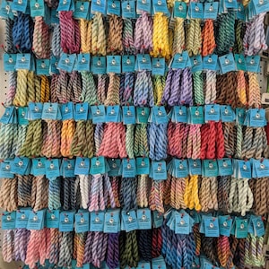 Puede incluir: Una pared de exposición de ovillos de hilo de bordar de colores, cada uno con una etiqueta azul que dice "DMC Color Card". El hilo está dispuesto en filas y columnas, creando una exposición vibrante y organizada.
