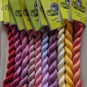 Op de afbeelding: Assortiment van 10 spoelen borduurgaren in verschillende tinten roze, rood, geel en oranje. De spoelen zijn genummerd en dragen de merknaam "Chameleon Hand-Counted Embroidery Floss".