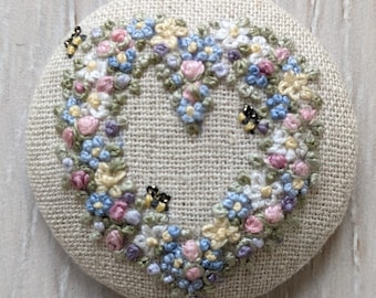 May include: A round, beige fabric pincushion with a heart-shaped design embroidered in shades of blue, pink, and yellow. The heart is surrounded by small, embroidered bees.