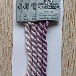 Peut inclure: Cinq bobines de fil à broder en soie dégradé dans des tons de violet et de blanc. Les bobines sont étiquetées "Chameleon Hand Colored Embroidery Threads" et "Winelands Au Ver à Soie - Sole de Paris 100% pure stranded silk Imported from France" et "Shades of Africa".