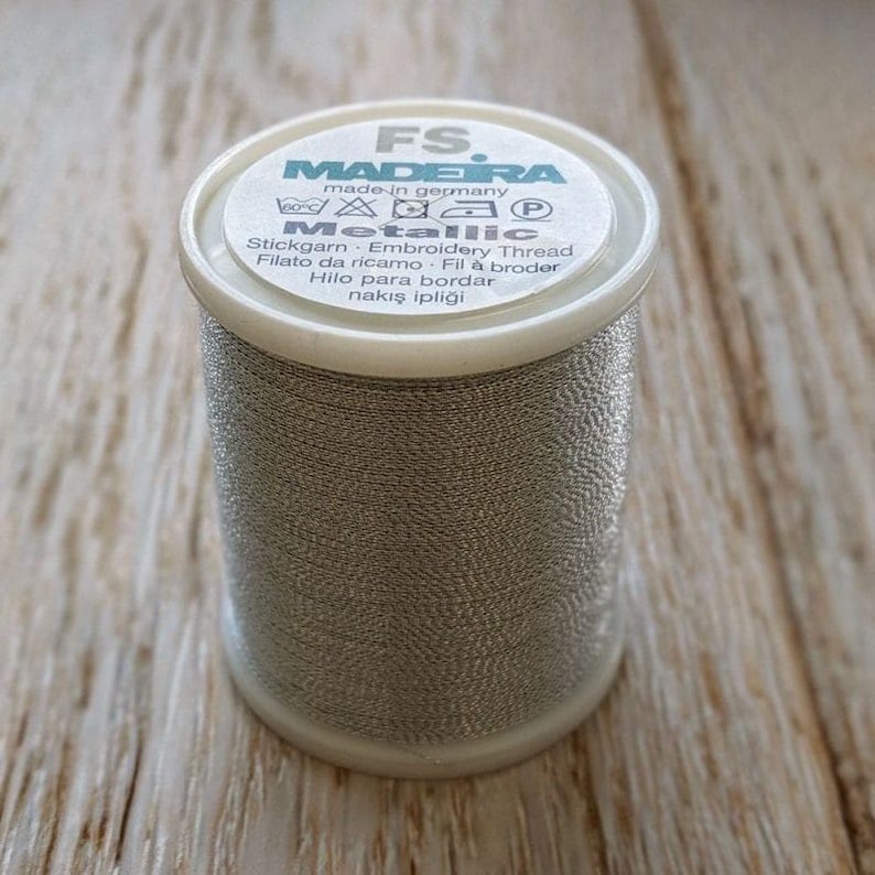 May include: A spool of silver metallic embroidery thread from Madeira, made in Germany. The label reads "FS. MADEIRA made in germany Metallic Stickgarn Embroidery Thread Filato da ricamo Fil &agrave; broder Hilo para bordar nakış ipliği".