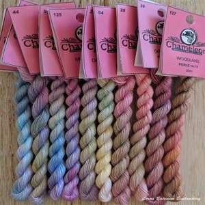 May include: Eight skeins of embroidery floss in a gradient of colours, from blue to pink. The floss is wound into balls and labelled with numbers, including 44, 9, 125, 64, 20, 39, and 127. The labels also include the text "Chameleon" and "Woodland Perle no. 12 20m".