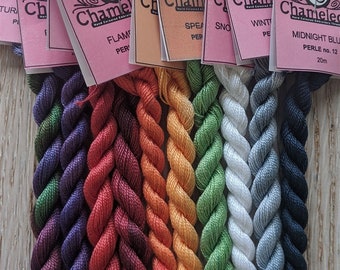 Puede incluir: Ocho carretes de hilo de bordar en varios colores, incluyendo morado, rojo, naranja, verde, blanco y azul. Cada carrete está etiquetado con el nombre de la marca "Chameleon" y un número. El hilo está enrollado alrededor de los carretes en un patrón retorcido.