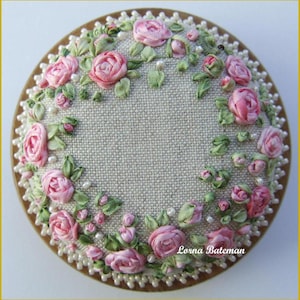 May include: A circular embroidery hoop with a beige linen fabric background. The fabric is decorated with a wreath of pink satin ribbon roses and green satin ribbon leaves. The wreath is accented with white pearl beads.