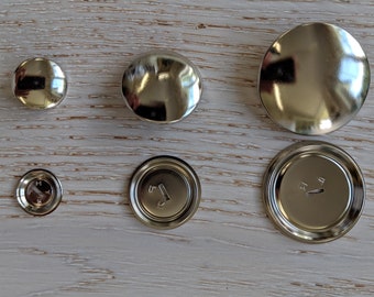Könnte beinhalten: Sortiment von silbernen Metallknöpfen in verschiedenen Größen. Die Knöpfe sind rund und haben eine glatte, glänzende Oberfläche. Einige Knöpfe haben eine kleine Vertiefung in der Mitte.