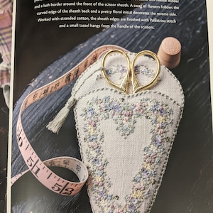 May include: A heart-shaped needle minder and scissor sheath embroidered with pastel flowers and a floral initial. The sheath is made of natural linen and has a small tassel hanging from the handle of the scissors.