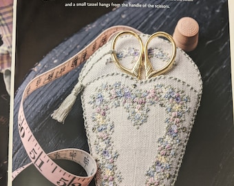 May include: A heart-shaped needle minder and scissor sheath embroidered with pastel flowers and a floral initial. The sheath is made of natural linen and has a small tassel hanging from the handle of the scissors.