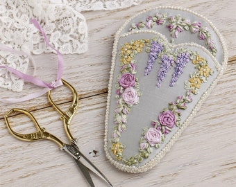 May include: A gray fabric pincushion with a floral design embroidered in shades of purple, pink, and yellow. The pincushion is surrounded by a row of white pearls. A pair of gold scissors is lying on a wooden surface.