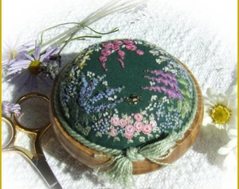 May include: A green fabric pincushion with embroidered flowers and a bee, surrounded by a wooden base with a green ribbon and tassel.