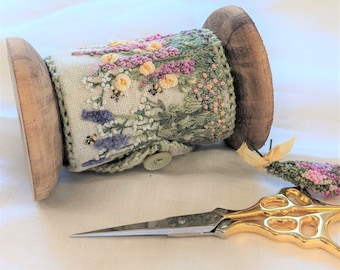 May include: A wooden spool with a hand-stitched floral design in shades of pink, purple, green, and yellow. The spool is wrapped with a white fabric and has a button on the side. A pair of gold scissors are also in the image.