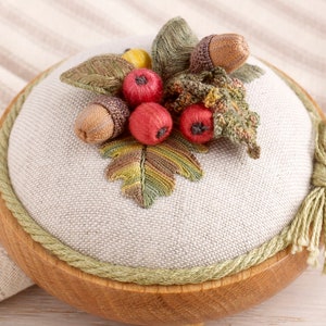 Puede incluir: Un cojín de alfileres redondo de madera con una parte superior de lino. La parte superior está decorada con un diseño bordado de hojas de otoño, bellotas y bayas en tonos de verde, marrón y rojo.