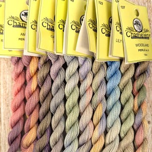 May include: A selection of Chameleon perle cotton threads in a range of colours. Each skein is labelled with a number and the brand name. The threads are in shades of pink, orange, green, blue, and purple. The labels also indicate the thread is 16m in length.