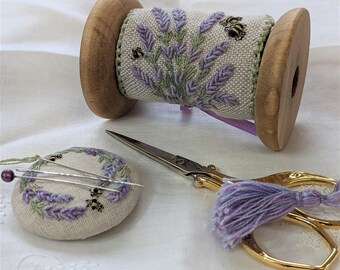 May include: A wooden spool with a lavender and bee embroidery design, a small lavender pincushion with pins, and gold scissors with a lavender tassel.