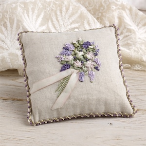 Puede incluir: Una pequeña almohada cuadrada de lino con un ramo de flores de cinta rosa y morada cosido a mano. La almohada tiene un ribete de cuentas doradas y moradas.