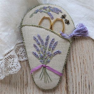 May include: A beige fabric scissor holder with a lavender embroidery design. The holder has a pocket for scissors and a tassel.