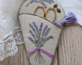 May include: A beige fabric scissor holder with a lavender embroidery design. The holder has a pocket for scissors and a tassel.