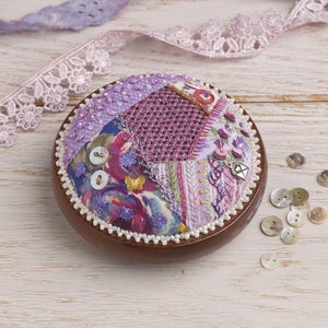 May include: A round, purple and white patchwork pincushion with a wooden base and pearl trim. The pincushion is filled with buttons and beads.