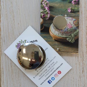 Puede incluir: Un imán redondo plateado para agujas con tapa, sobre una tarjeta blanca con texto y un diseño floral. La tarjeta está sobre una superficie de madera, cerca de un alfiletero y flores.