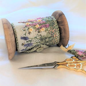 Puede incluir: Un carrete de cinta bordada con un diseño floral con flores rosas, moradas, amarillas y verdes. La cinta está envuelta alrededor de un carrete de madera y un par de tijeras doradas están sobre una superficie blanca.